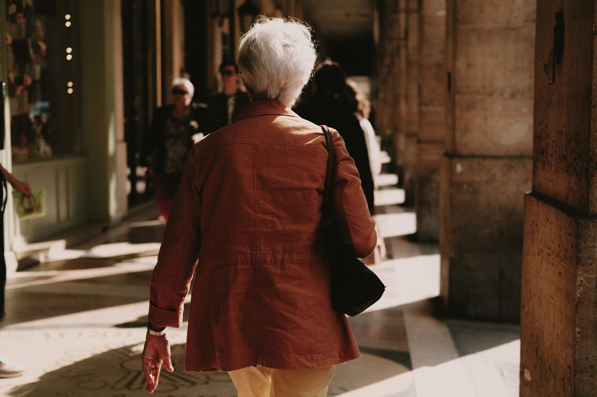 Une femme âgée en manteau rouge marche sous des arcades.